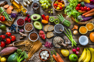 grand groupe de fruits frais multicolores, légumes, céréales et épices tourné d'en haut sur fond de bois. La composition comprend pomme verte, kiwi, poire, grenade, orange, noix de coco, banane, raisin, baies, gingembre, amandes, pistache, huile d'olive, olives, baies de goji, graines de chia, haricots pinto, muscade, romarin, radis, tomates, carotte, chou frisé, avocat, oignon, riz, cacao en poudre, patate douce, pâtes complètes, tofu, laitue, maïs, brocoli, poivron, asperges, haricots verts, entre autres.