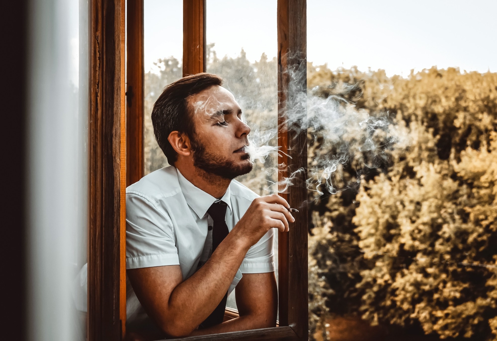 A man smokes a cigarette on the balcony against the background of autumn trees with golden foliage.