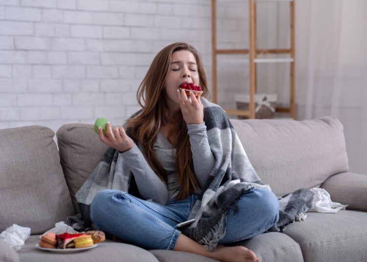 Sad young lady eating sweets on sofa at home