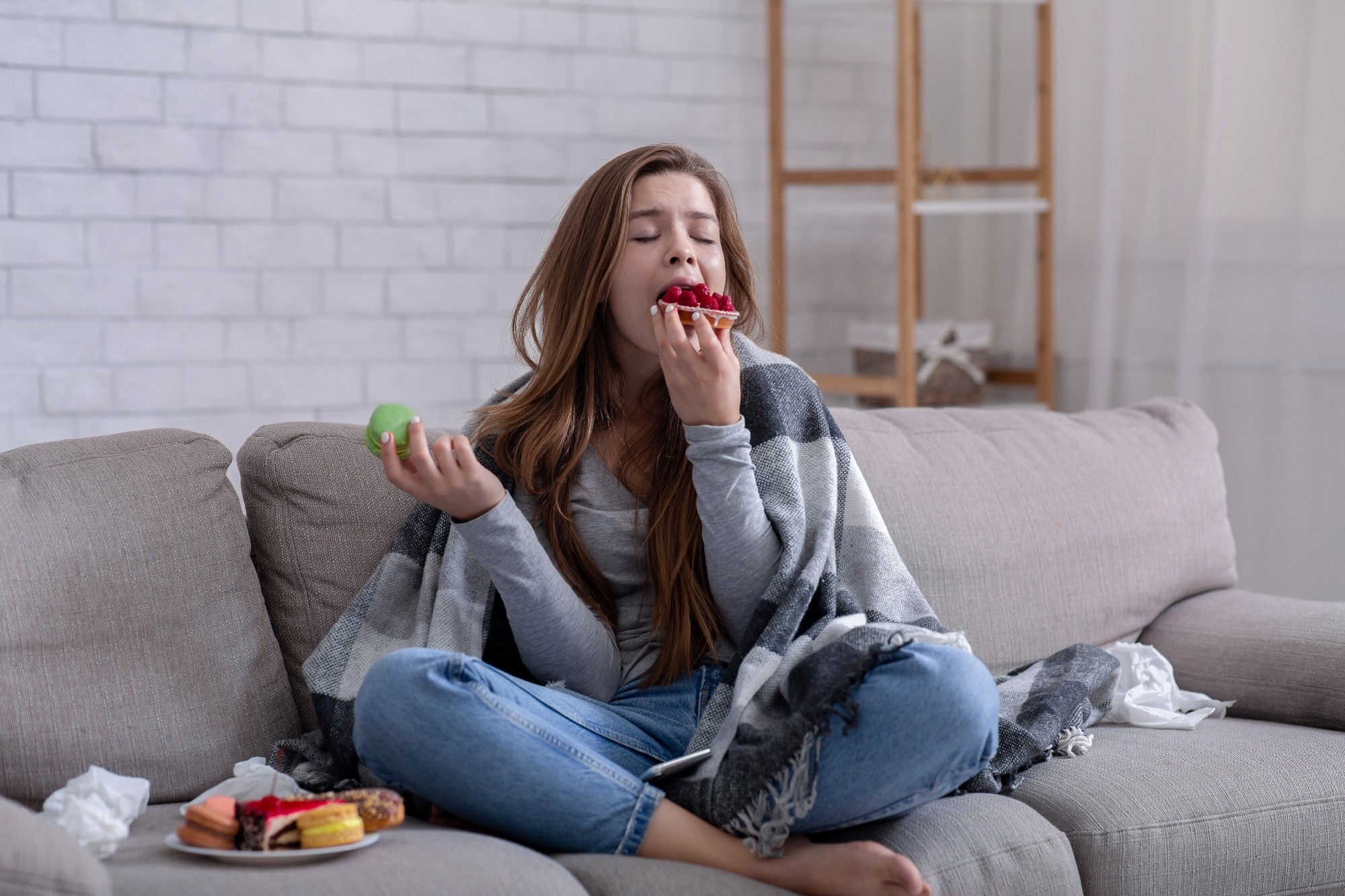 Sad young lady eating sweets on sofa at home