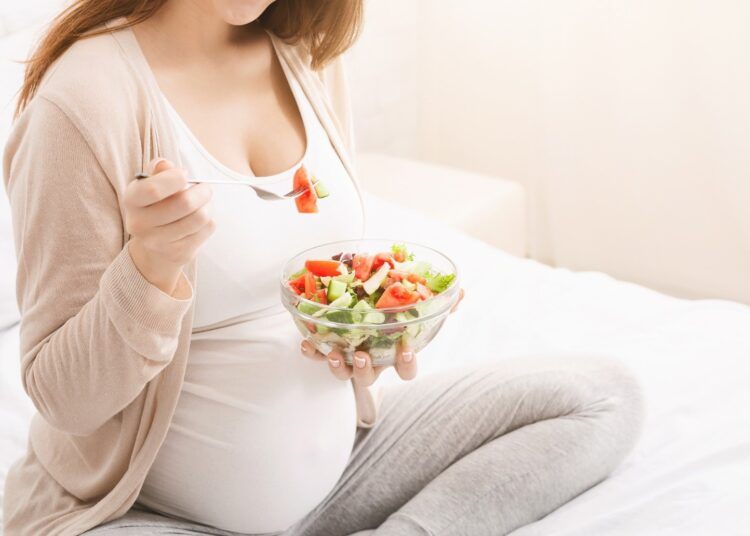 A pregnant woman is sitting on a bed, holding a bowl of salad in her hands