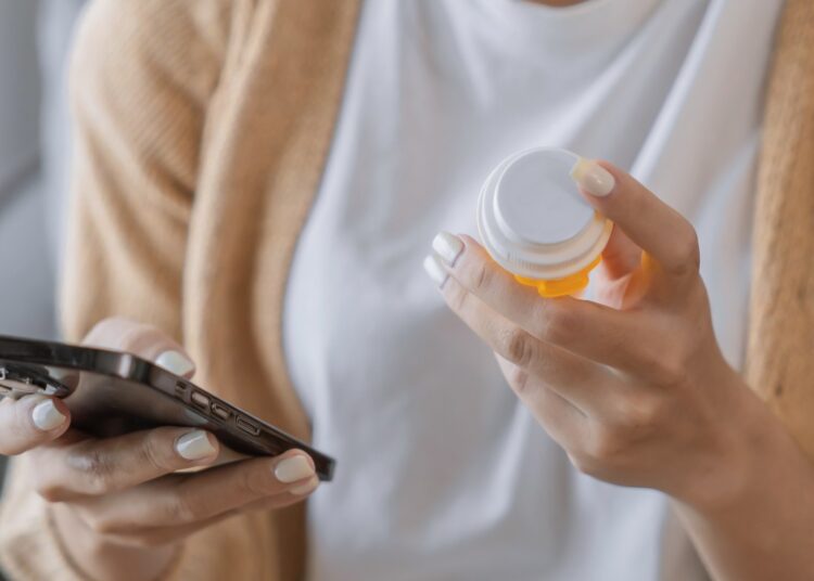 Woman reading information online on mobile phone holding bottled prescription drug