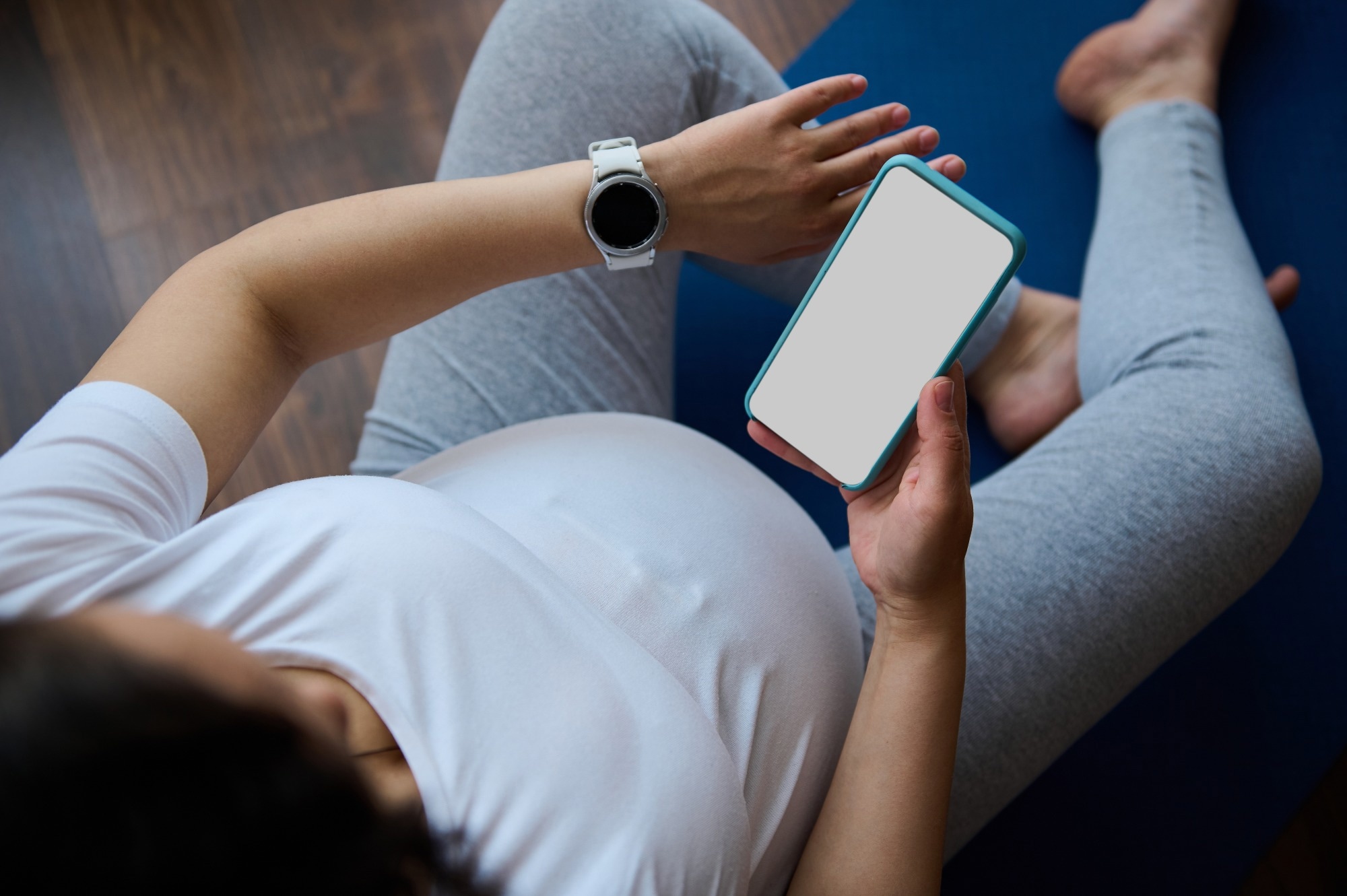 Top view pregnant woman checks smart watch to control pulse and heart rate, relaxing with smartphone
