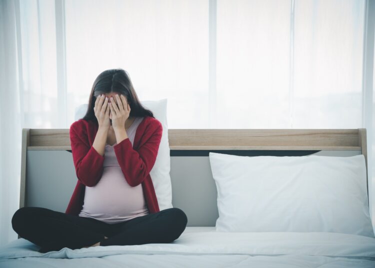 Pregnant woman in casual home outfit sitting on bed and feeling upset and sad