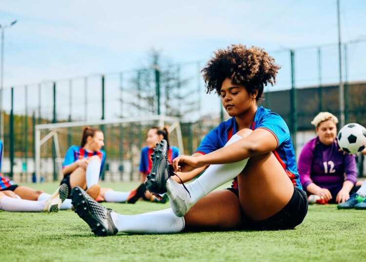 Young female footballer sat on pitch putting on training shoes