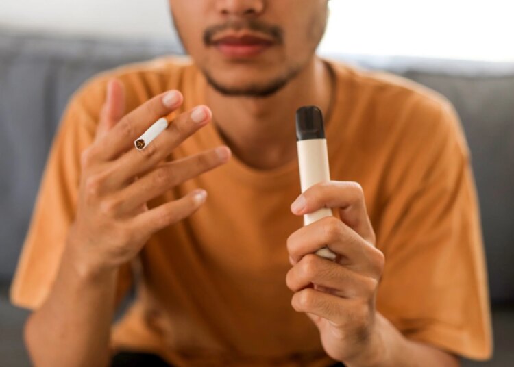 Young Asian man holding a cigarette and e-cigarette vaping device while sitting in couch