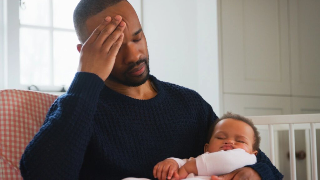 Stressed New Father Sitting In Chair Holding Sleeping Baby Girl In Nursery At Home