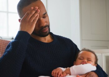 Stressed New Father Sitting In Chair Holding Sleeping Baby Girl In Nursery At Home
