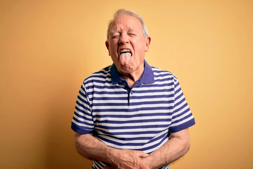 Grey haired senior man wearing casual navy striped t-shirt standing over yellow background sticking tongue out happy with funny expression.