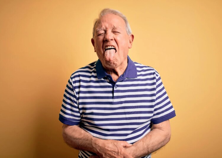 Grey haired senior man wearing casual navy striped t-shirt standing over yellow background sticking tongue out happy with funny expression.