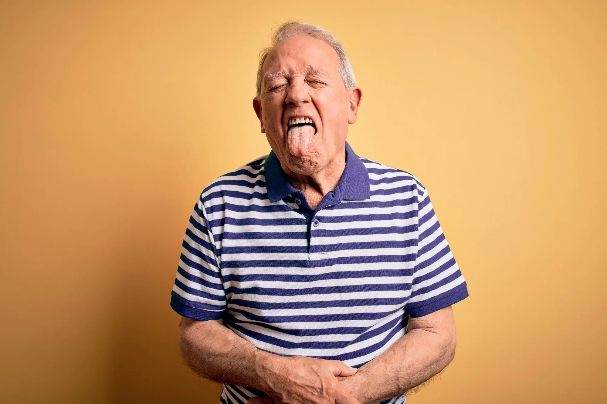 Grey haired senior man wearing casual navy striped t-shirt standing over yellow background sticking tongue out happy with funny expression.