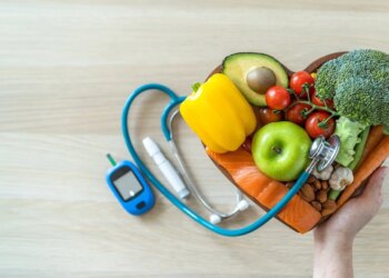 A hand holds a heart-shaped bowl filled with fresh healthy foods - including vegetables, fruit, fish, and nuts - next to a glucose meter and stethoscope, symbolizing diet and heart health management in diabetes.