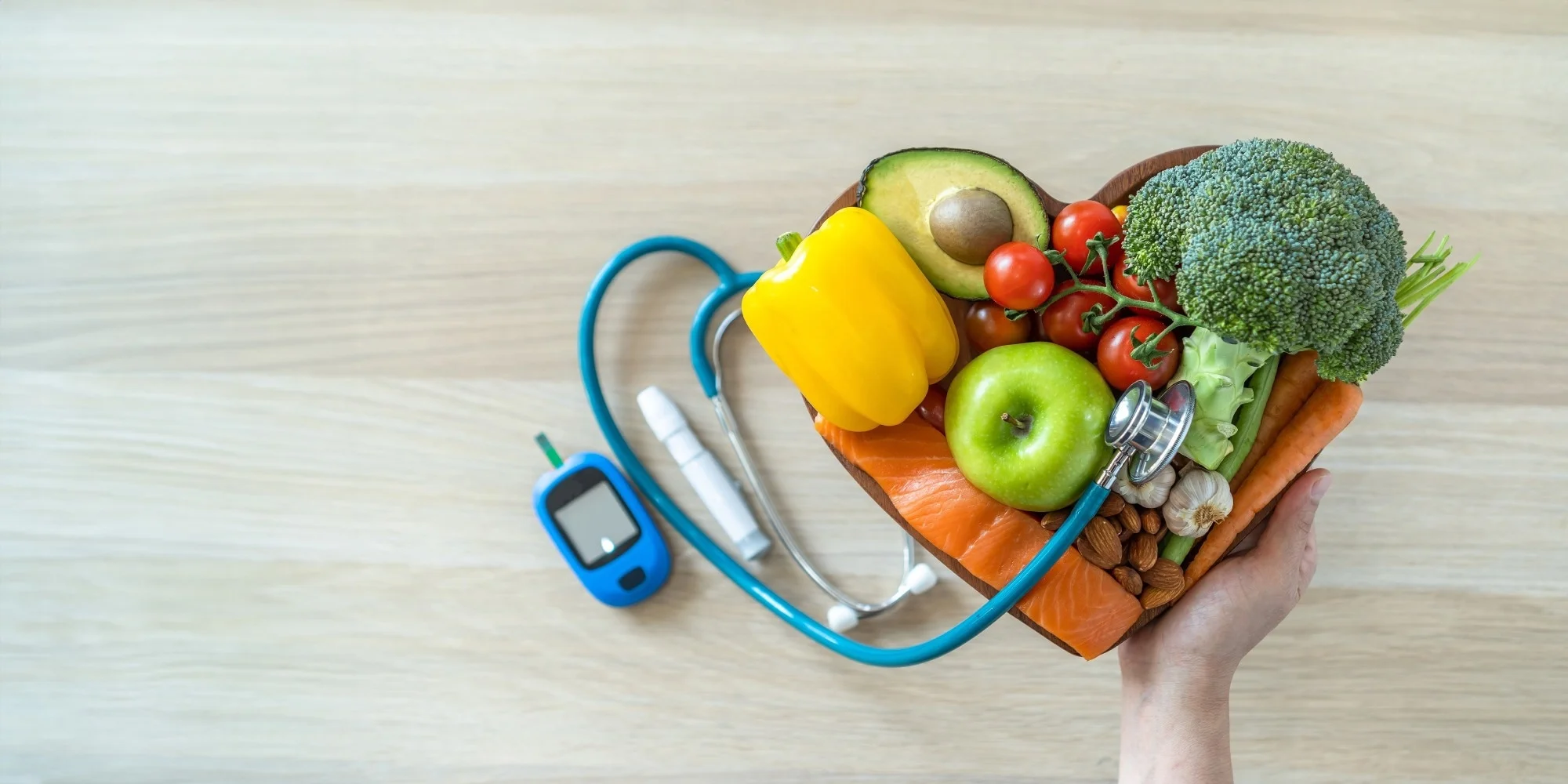 A hand holds a heart-shaped bowl filled with fresh healthy foods - including vegetables, fruit, fish, and nuts - next to a glucose meter and stethoscope, symbolizing diet and heart health management in diabetes.