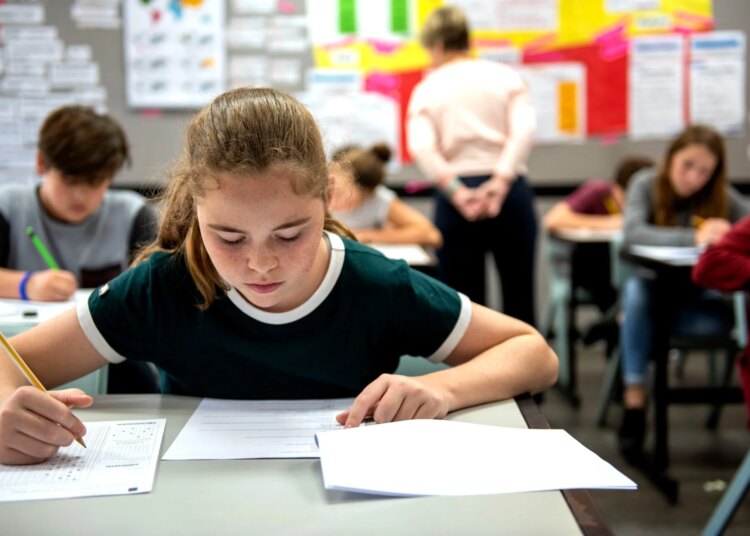 Students in a classroom taking a test, focused on their papers. A teacher observes in the background, ensuring a quiet environment.
