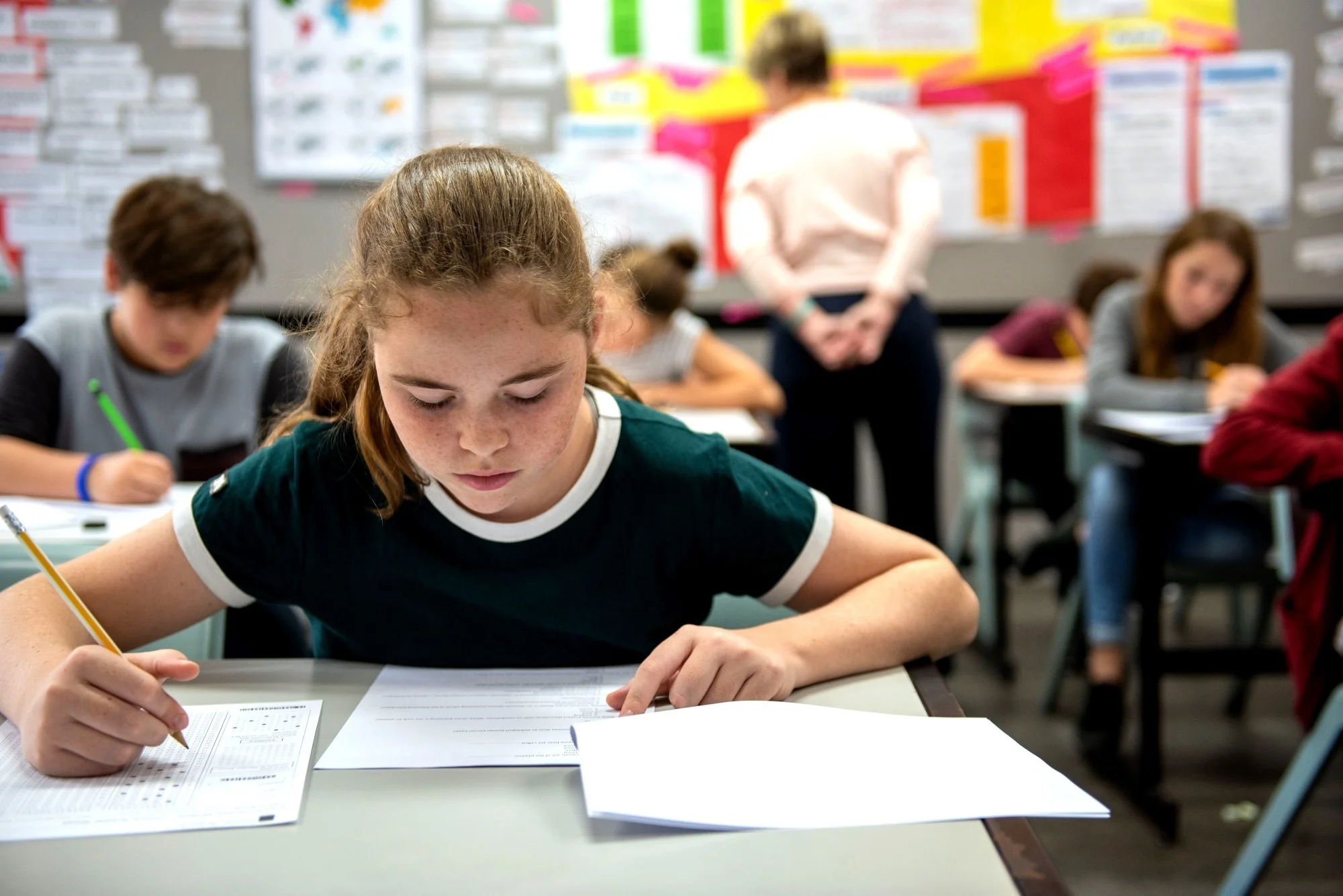 Students in a classroom taking a test, focused on their papers. A teacher observes in the background, ensuring a quiet environment.