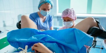 Female gynecologist with young woman assistant in mask and hair cap with unrecognizable patient in gynecology chair. Hysteroscopy operation