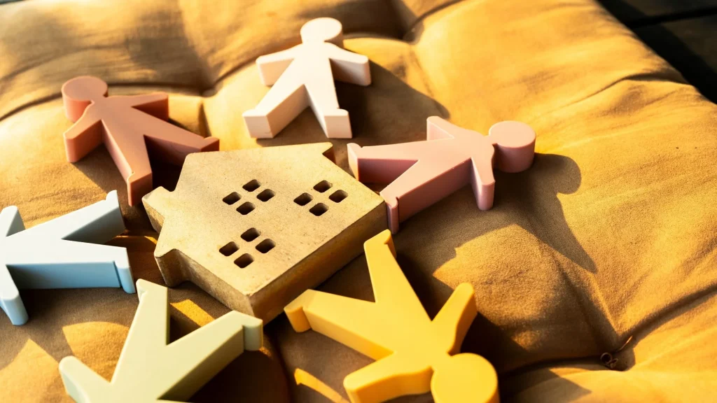 Colorful human figures surrounding a wooden house symbol on a cozy cushion. Concept of community, family, diversity, home sharing, social unity, co-living, and inclusive housing.