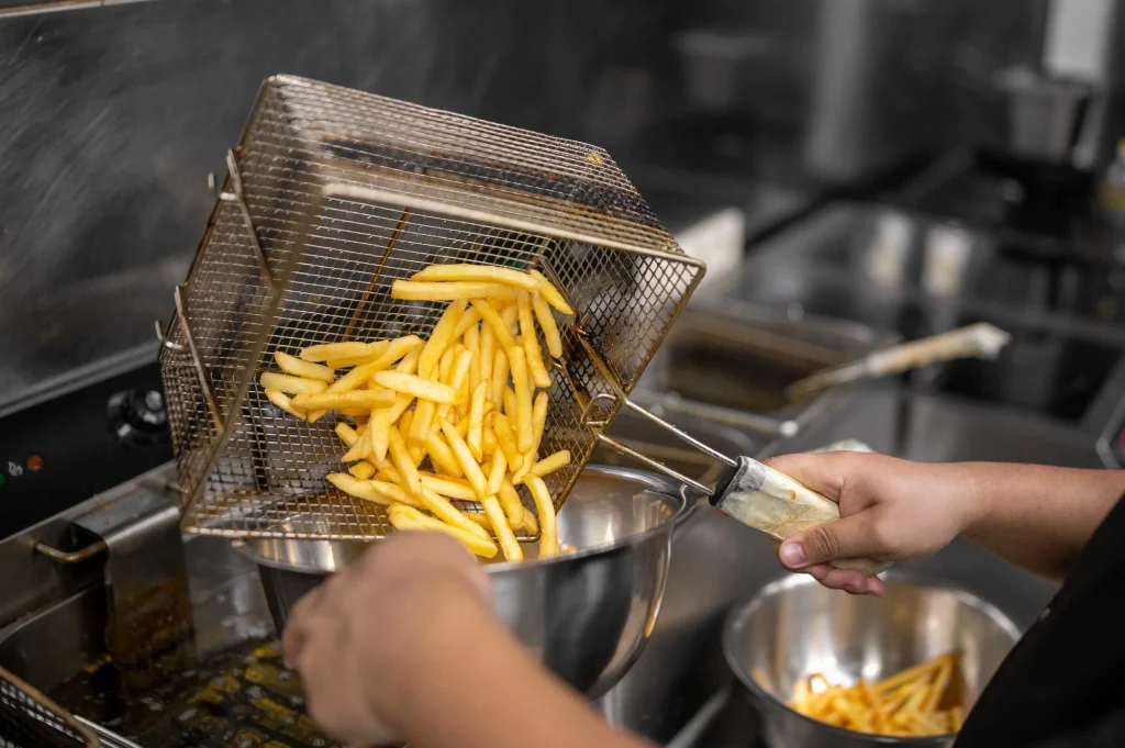 A chef empties fresh, golden french fries from a wire deep fryer basket into a metal bowl in a commercial kitchen.