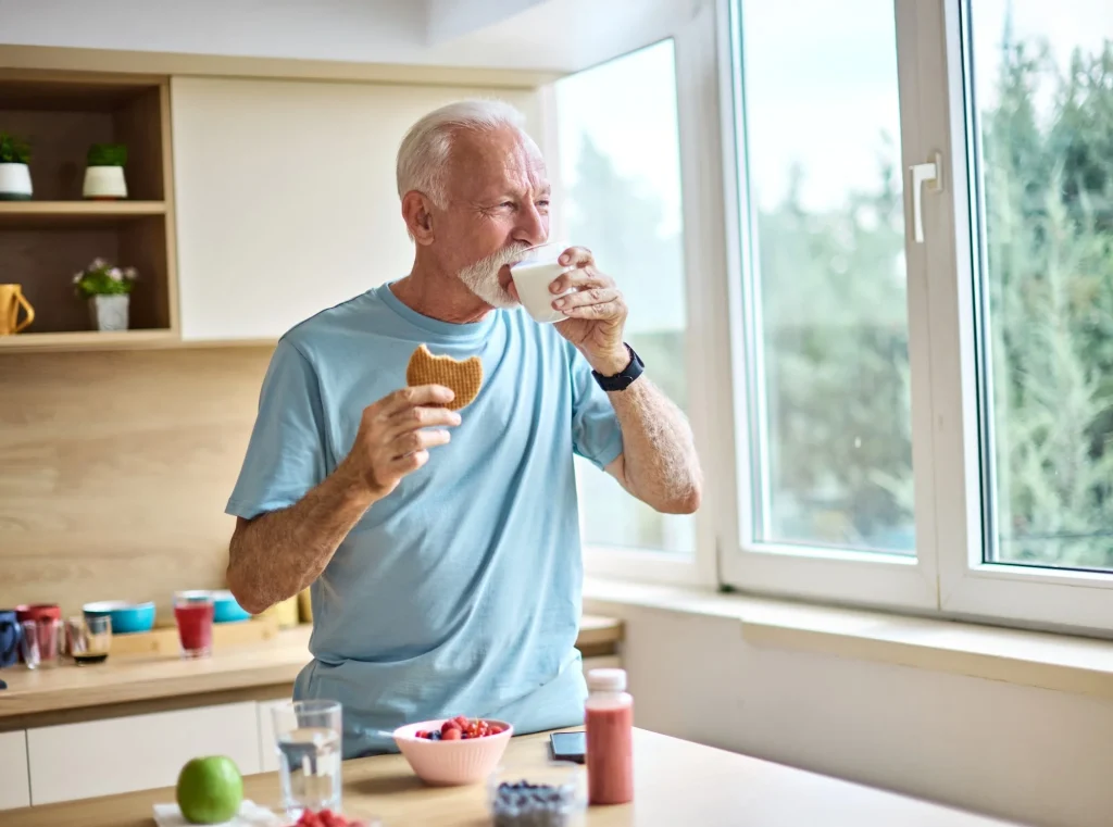 A senior man is eating a cokie energy bar and drinking milk in kitchen.
